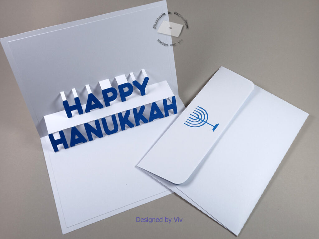 An open card with pop up words Happy Hanukkah and blue letters glued to the pop up element. An envelope with a nine candle candlestick cut out of the flap and blue card glued behind it.