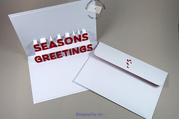 Card shown from an angle with the pop up letters Seasons Greetings. Red letters are glued to each individual pop up letter. An envelope with a candy stick cut out of the flap and red card glued behind it.