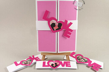 A gatefold card shown closed with a belly band with the word love diagonally across it. The O of the Love is replaced with a heart frame where a photo can be placed. Three other variations of the belly band and the Love photo frame lie in front.